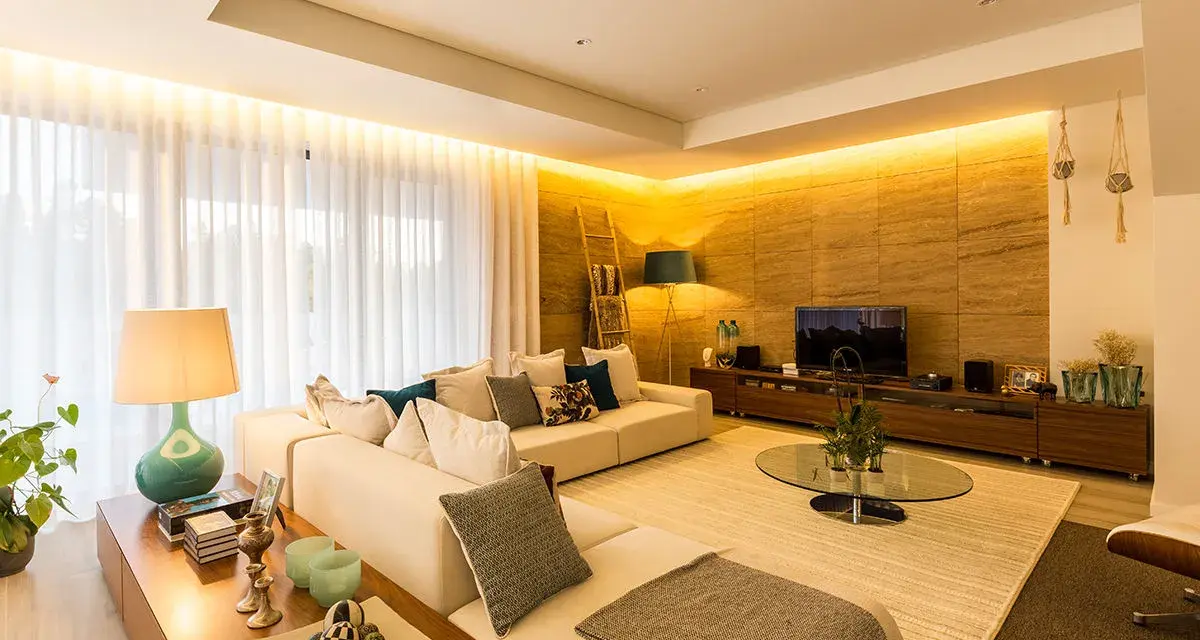 Salón moderno con sofá blanco, mesa de centro de cristal y **luz indirecta** que ilumina la pared de piedra y las cortinas.