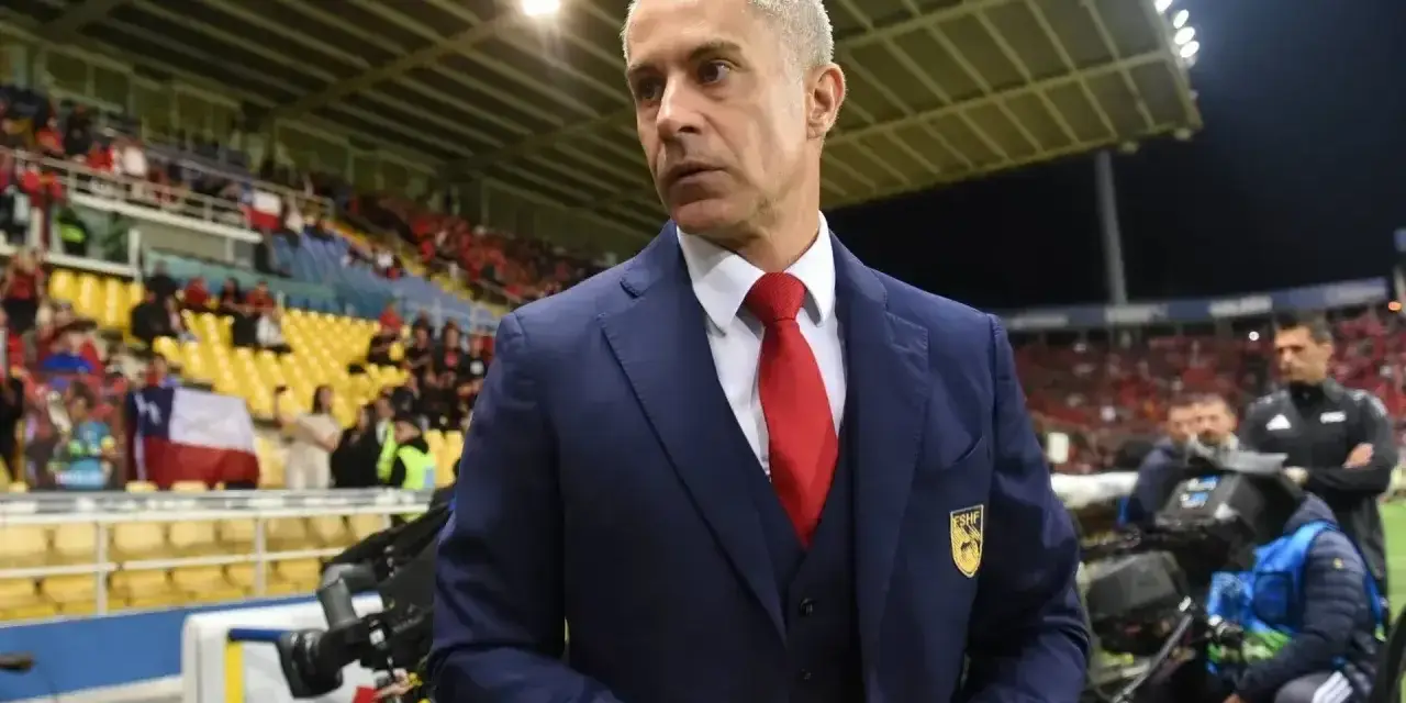 Sylvinho, trener w granatowym garniturze i czerwonym krawacie, stoi na tle trybun stadionu z flagą Chile.
