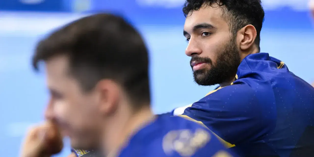 Hassan Kaddah, z brodą i kręconymi włosami, w niebieskiej bluzie z żółtymi detalami, spogląda w bok.