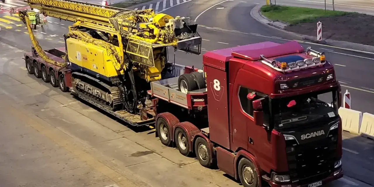 Czerwona ciężarówka Scania z przyczepą transportuje żółtą maszynę wiertniczą. Pilot pojazdu nienormatywnego czeka na przejazd.