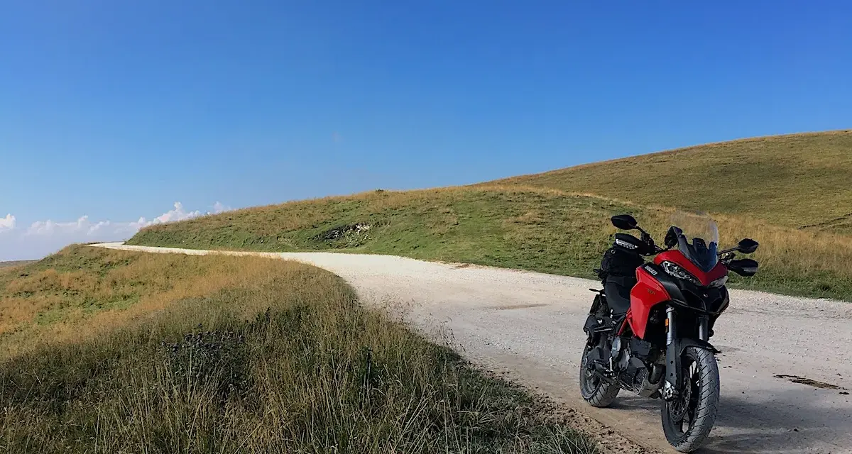 Ducati rossa ferma su strada sterrata, con un passo branchetto sullo sfondo e cielo azzurro.