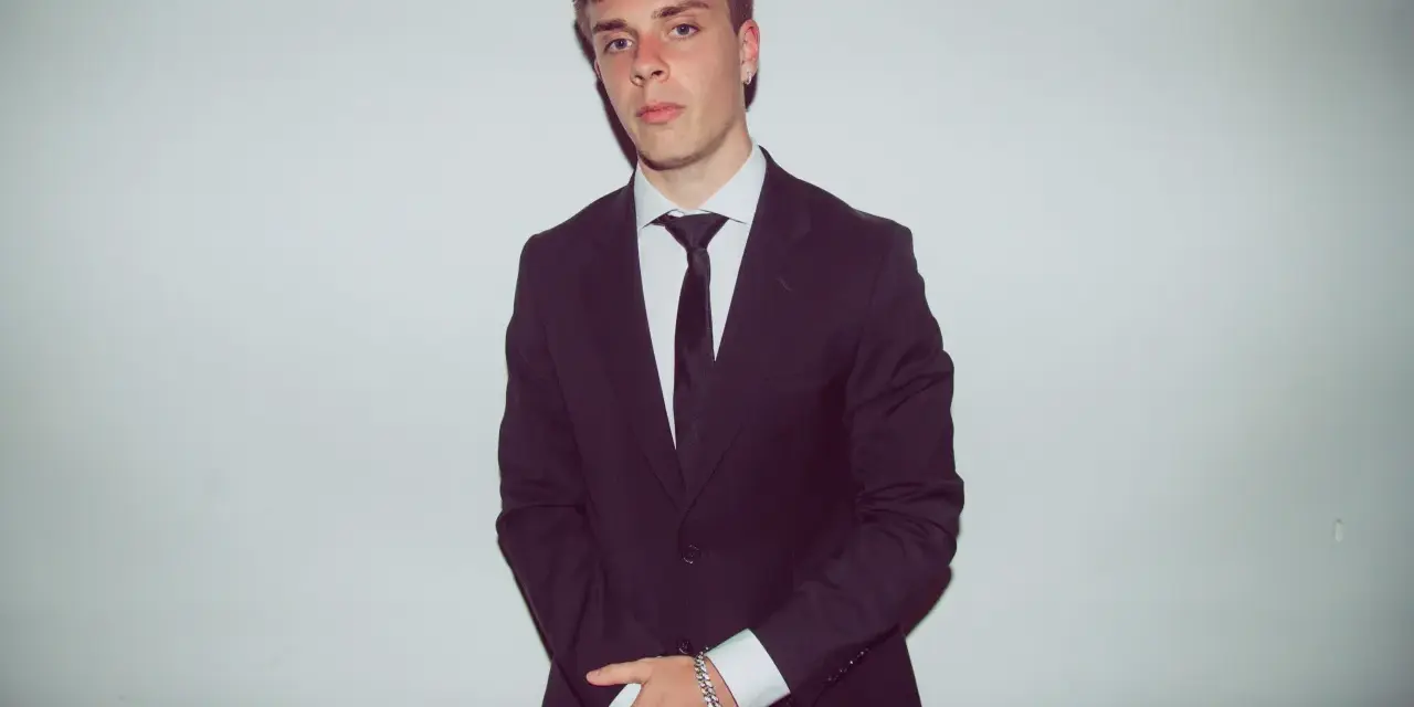 Młody mężczyzna w czarnym garniturze i białej koszuli, z krawatem. Kinny Zimner prezentuje się elegancko.