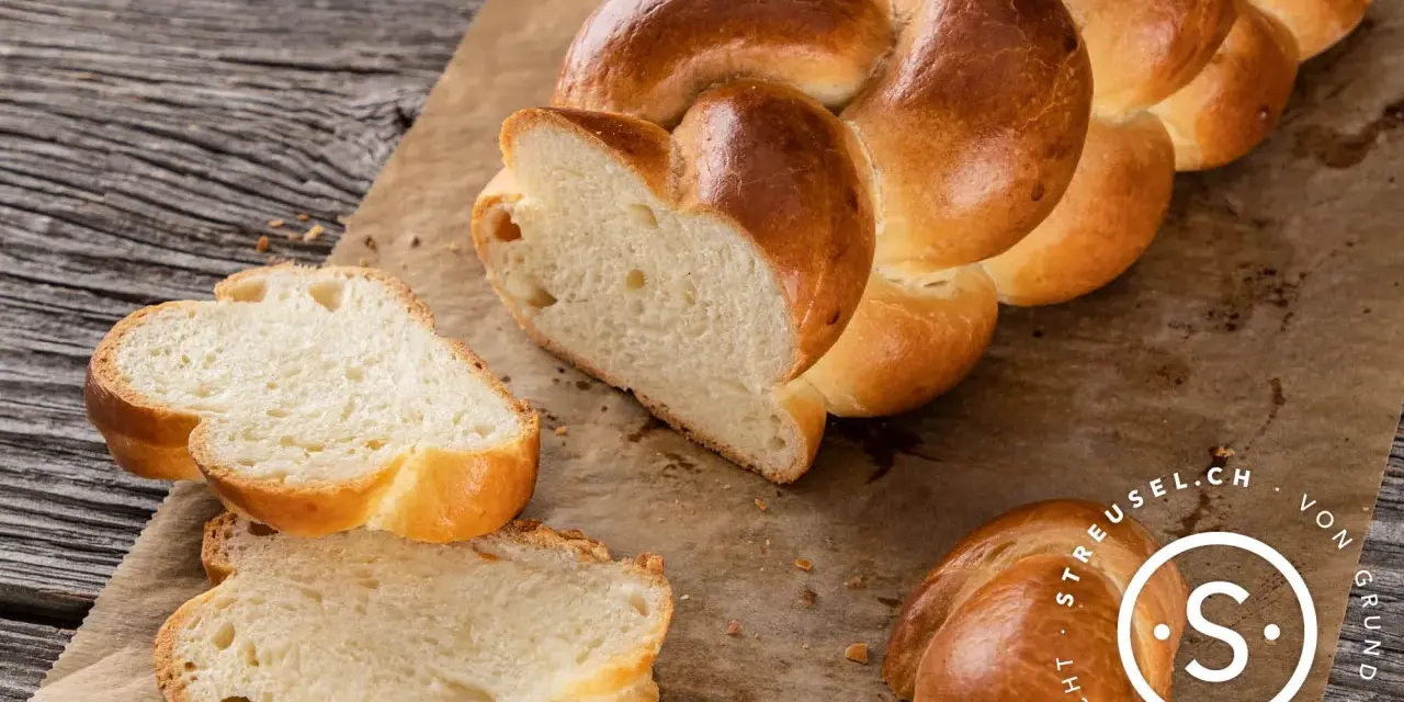 Ein goldbraunes Zopf Brot, frisch gebacken, liegt auf Backpapier. Zwei Scheiben sind abgetrennt und zeigen die weiche Krume.