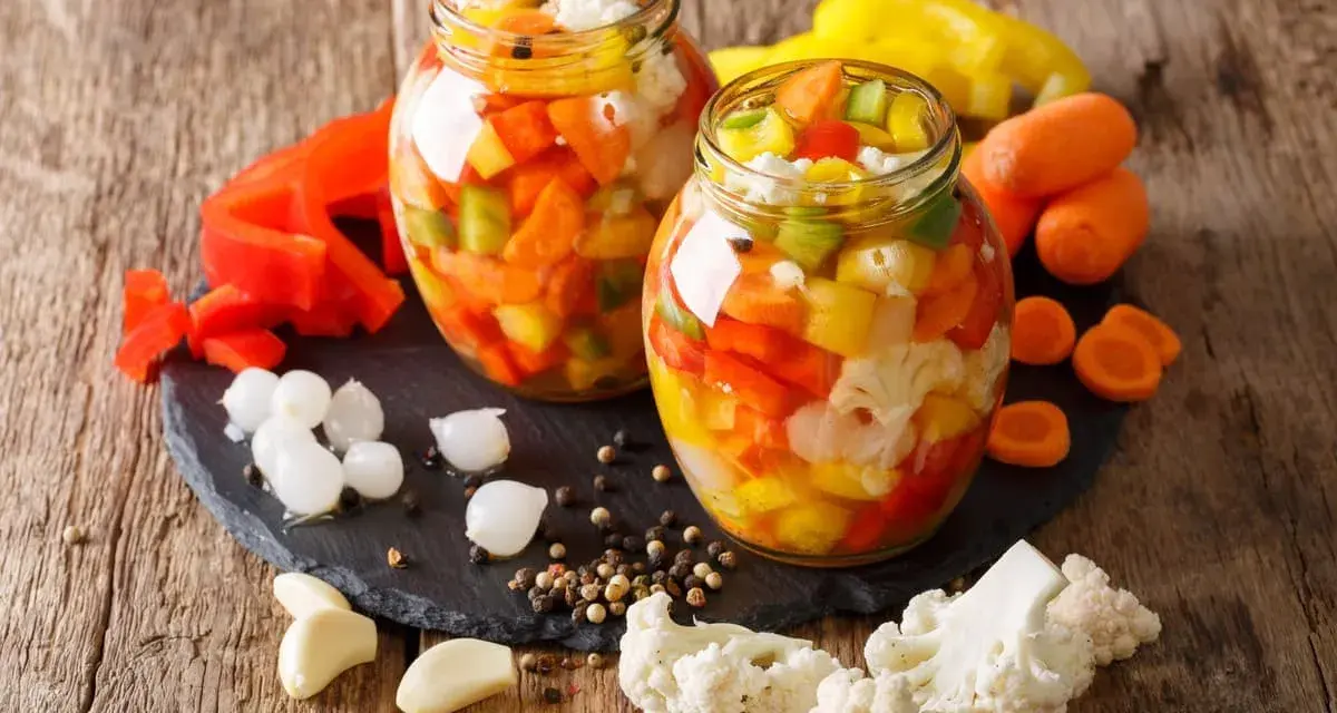 Due barattoli di giardiniera fatta in casa, pieni di verdure colorate come peperoni, carote e cavolfiore, su un tagliere di ardesia con ingredienti freschi intorno.