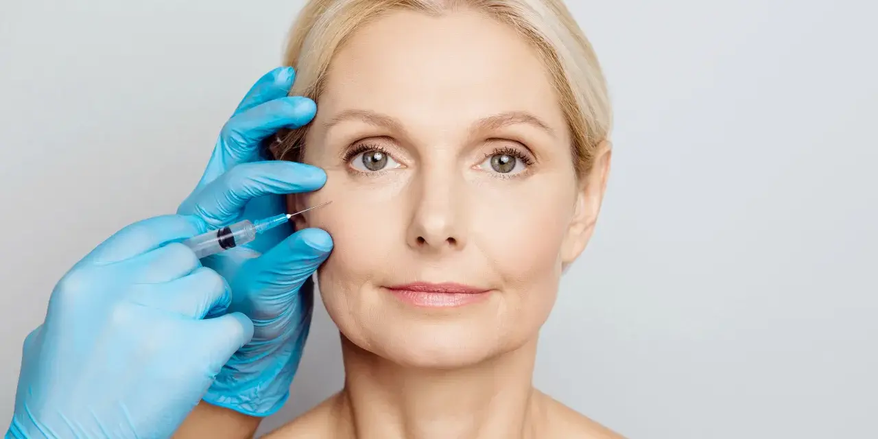 Zabieg z użyciem botoksu, czy botox jest bezpieczny? Dłoń w niebieskiej rękawiczce wykonuje zastrzyk w okolicę oka kobiety.