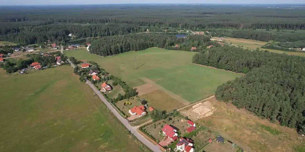 Widok z lotu ptaka na wieś otoczoną lasem. Widać domy z czerwonymi dachami, pola uprawne i jezioro. Każdy dom zajmuje swój kawałek ziemi, a całość tworzy malowniczy krajobraz.
