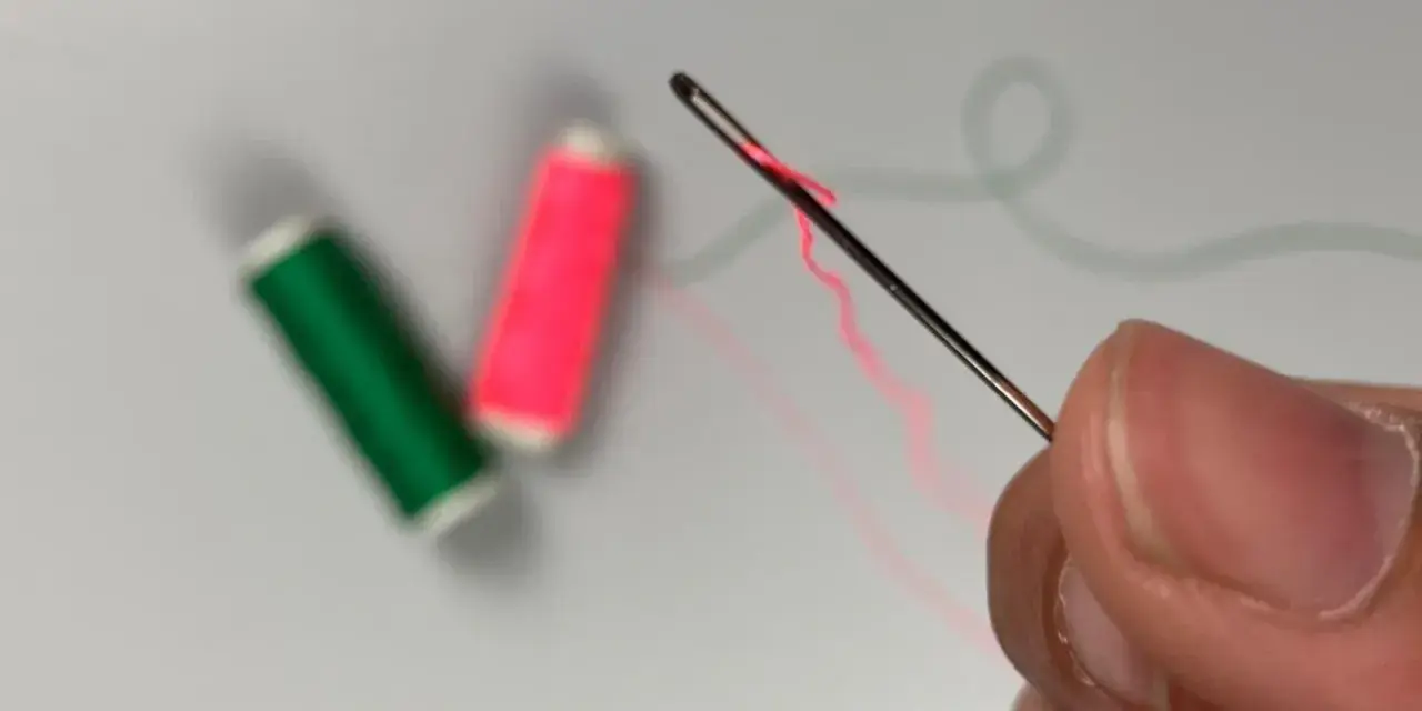 Dedos sujetan una aguja para meter hilos, con carretes de hilo verde y rosa desenfocados al fondo.