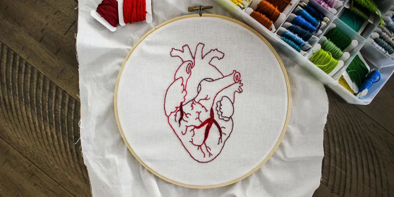 Hobbyści uczą się, jak wyszywać na ubraniach, tworząc anatomiczne serce na tamborku. Wokół kolorowe nici.