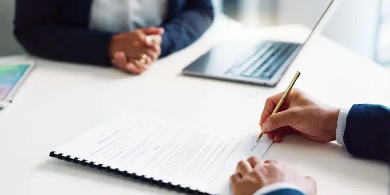 Persona firma un contrato de alquiler de habitación. A su lado, otra persona espera, con un portátil y una tablet sobre la mesa.