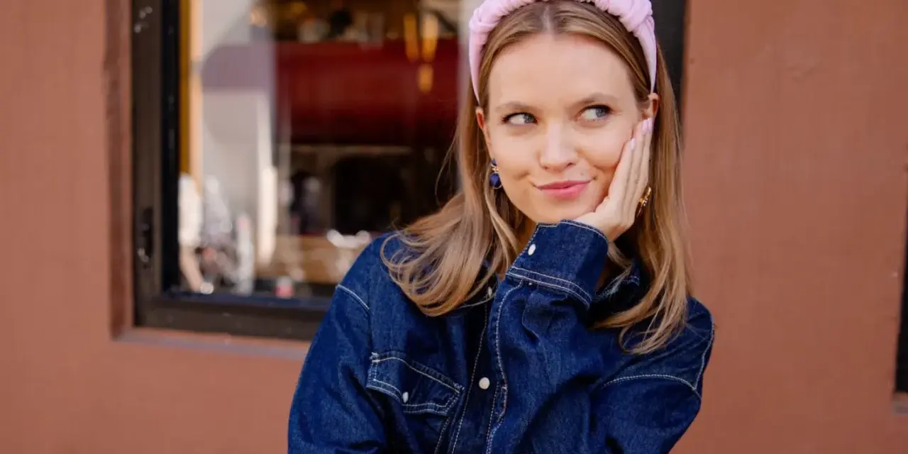 Aesthetic girl w jeansowej kurtce i różowej opasce na głowie, opiera policzek na dłoni, patrząc w bok.
