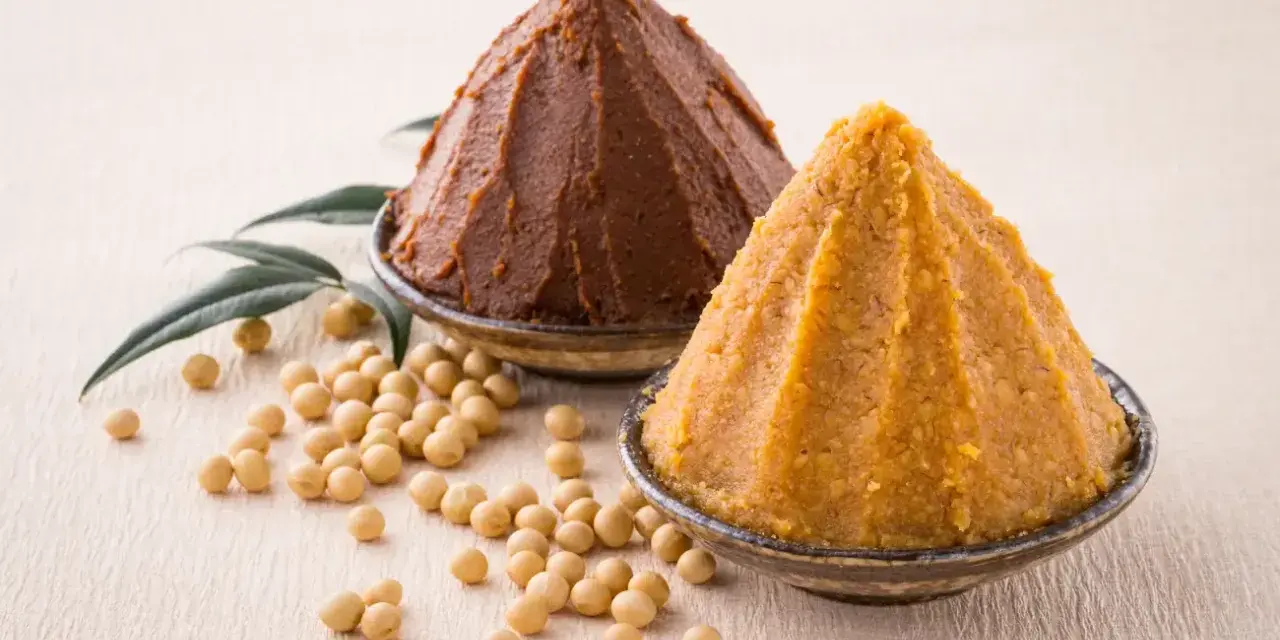 Dwie góry pasty miso, ciemna i jasna, obok ziaren soi i liści. Poznaj składniki do przygotowania tej aromatycznej pasty miso.