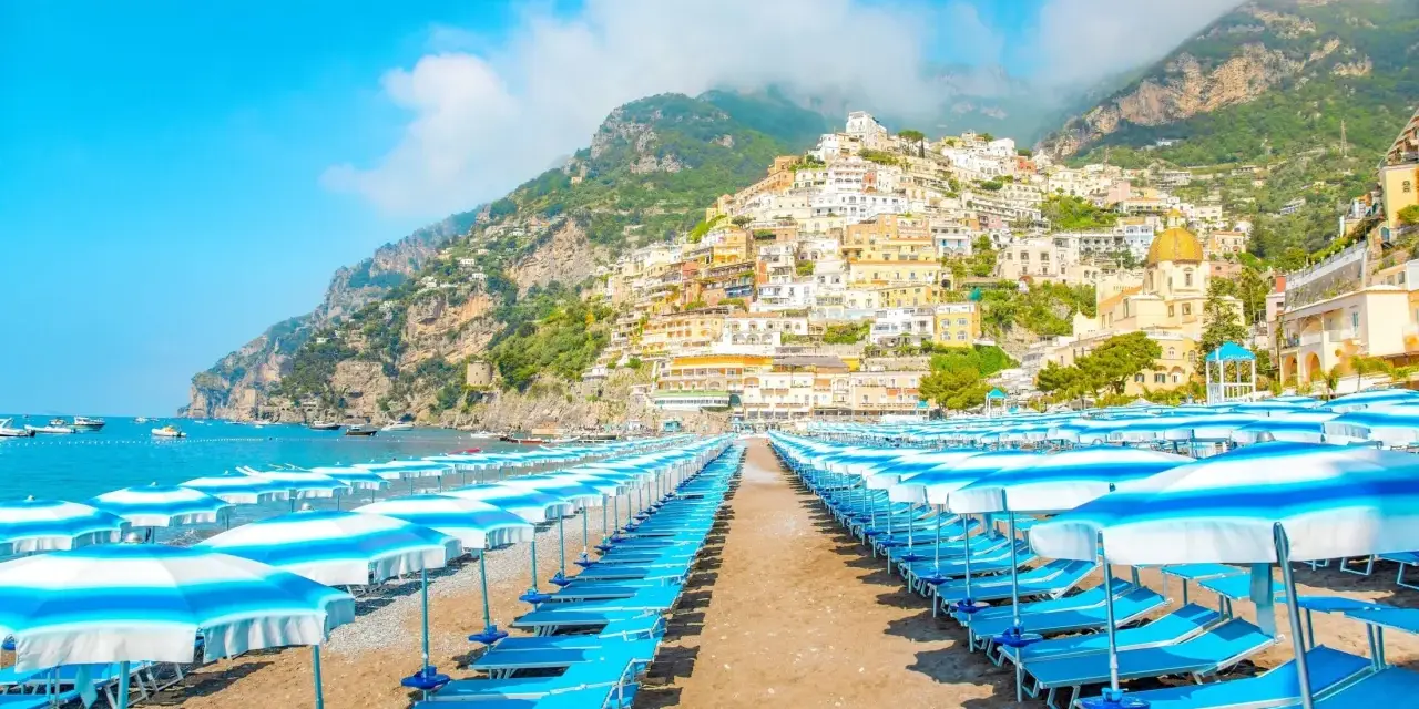 Rzędy leżaków i parasoli na plaży w Positano, Włochy. W tle widać malownicze miasteczko na zboczu góry i błękitne morze.