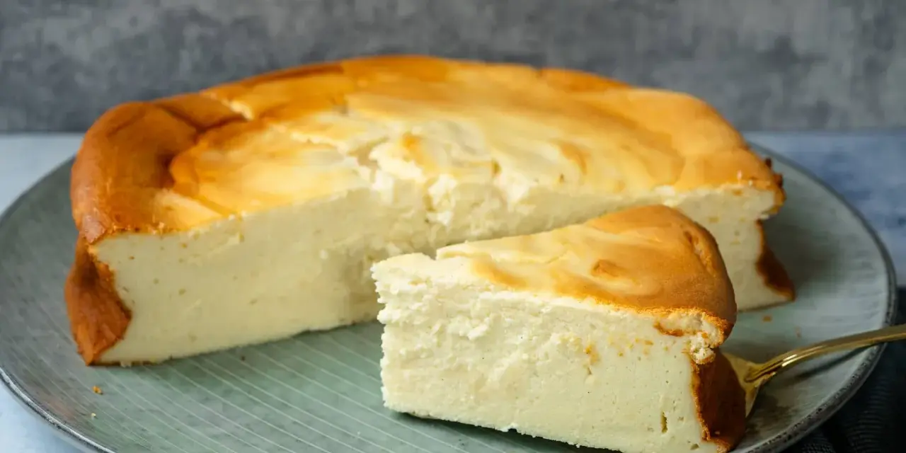Ein saftiger Quarkkuchen ohne Boden, goldbraun gebacken, liegt auf einem Teller. Ein Stück ist bereits angeschnitten und zeigt die cremige Konsistenz.