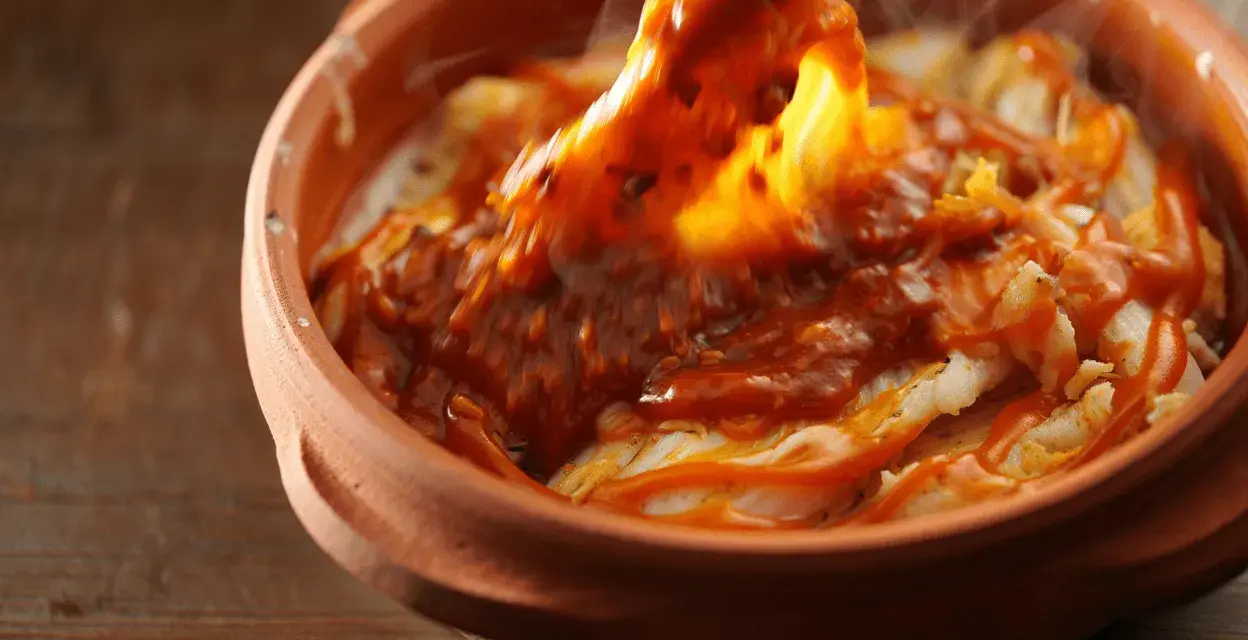 Cocochas de bacalao cubiertas con una salsa roja y burbujeante en una cazuela de barro.