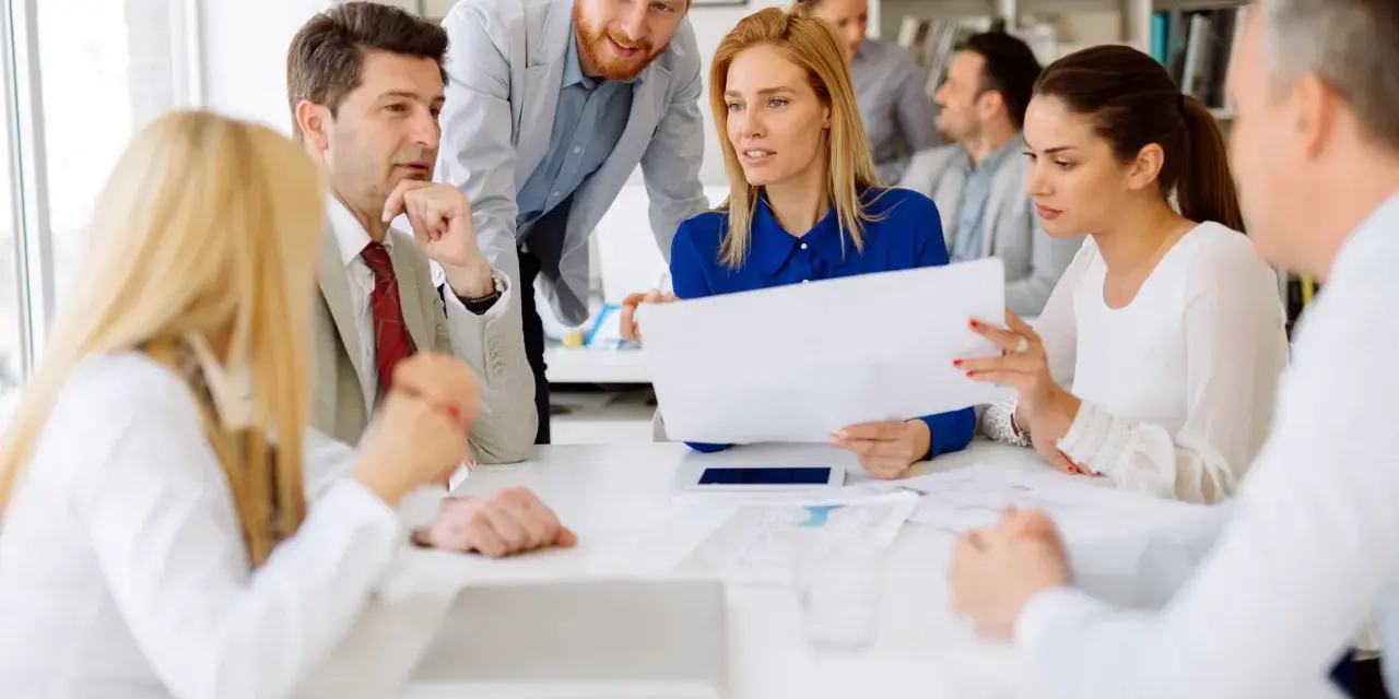 A diverse group of professionals discusses documents, possibly during board of directors recruitment or a strategic meeting.