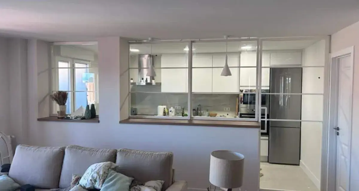 Un salón moderno con sofá y lámpara, separado de una cocina blanca y luminosa por una cristalera.
