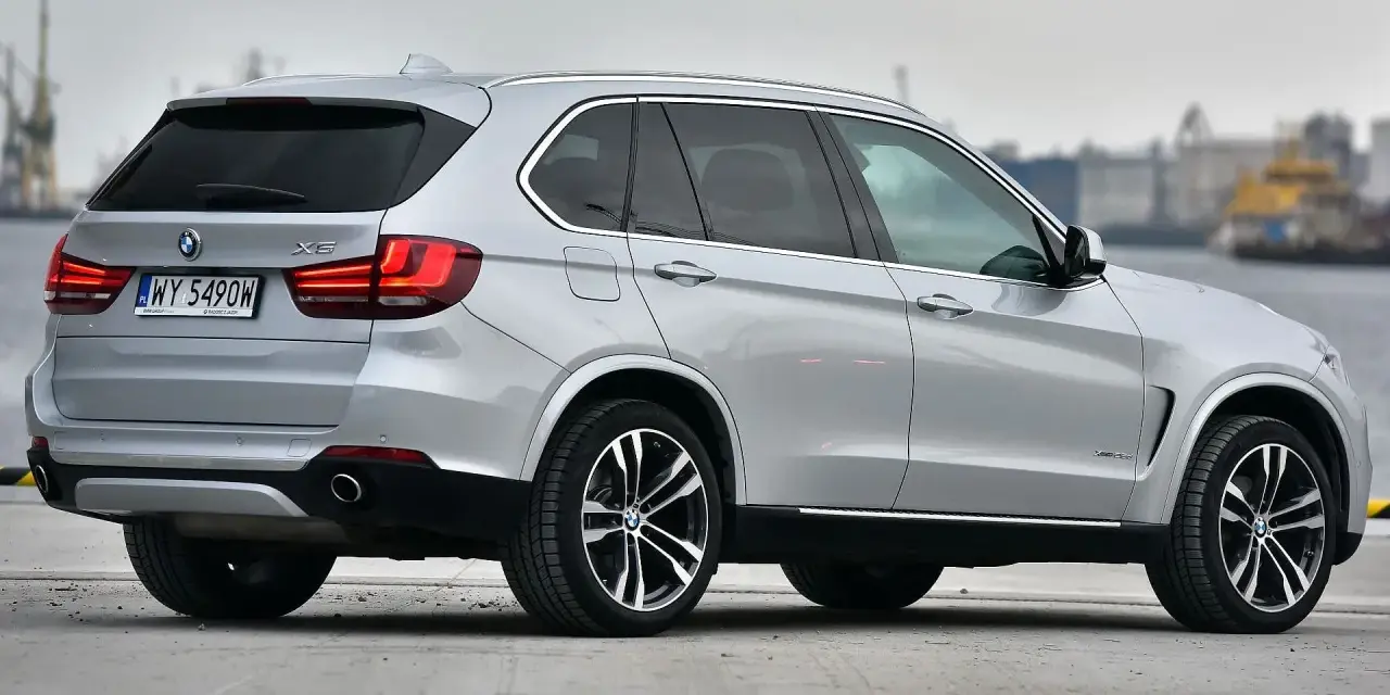 Srebrny BMW X5 z polskimi tablicami rejestracyjnymi stoi na betonowej nawierzchni, w tle widać port.