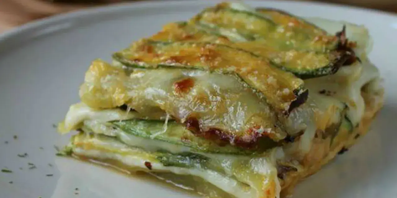 Una fetta di parmigiana di zucchine, stratificata con formaggio filante e zucchine grigliate, servita su un piatto bianco.