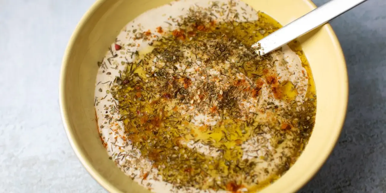 Cremiger Hummus Aufstrich in gelber Schüssel, beträufelt mit Olivenöl und bestreut mit Kräutern und Paprika.