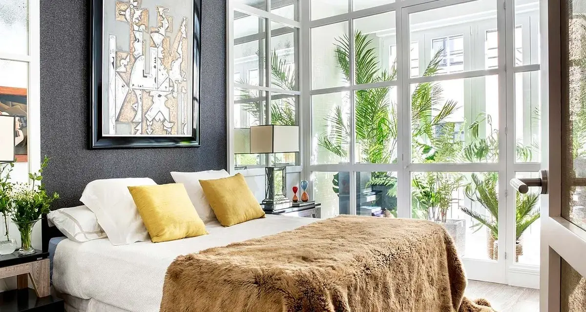 Dormitorios cálidos y modernos con cama mullida, mantas de pelo y gran ventanal con plantas.