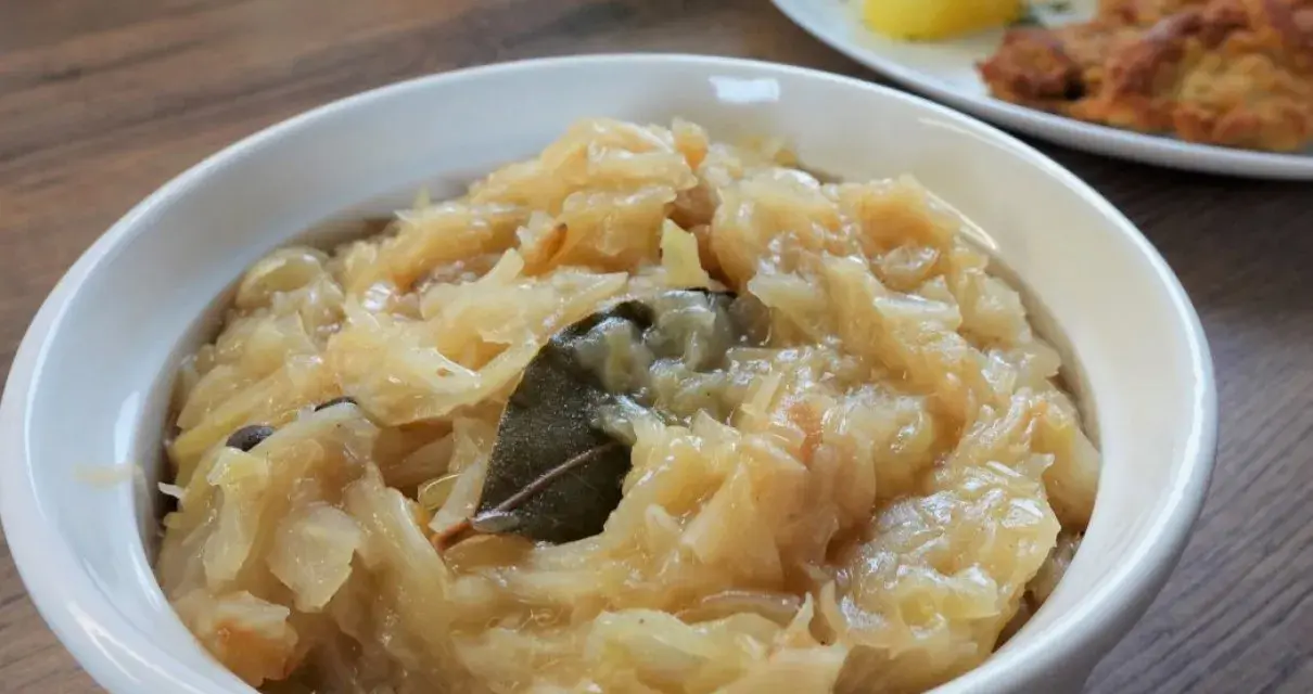 Pyszna, domowa kapusta zasmażana w białej miseczce, z liściem laurowym i ziarnami pieprzu. Idealny dodatek do obiadu.