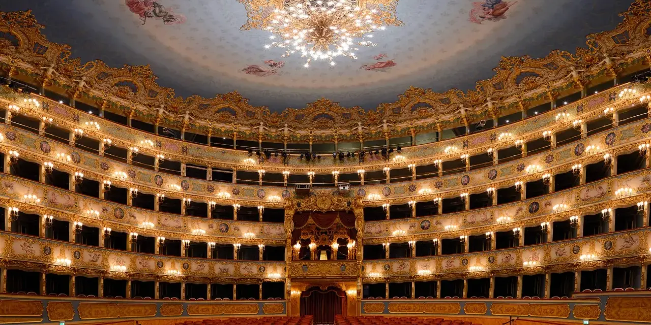 Interno sfarzoso del teatro La Fenice a Venezia, con palchi dorati, affreschi sul soffitto e un lampadario scintillante.