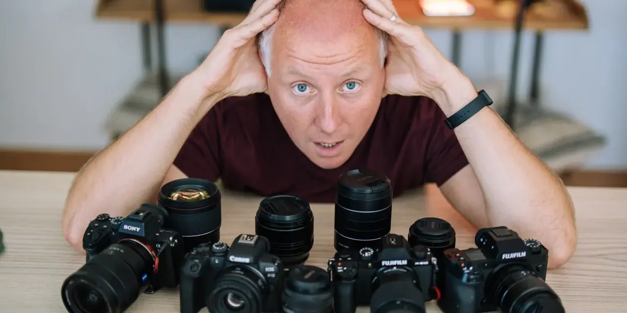 Mężczyzna z rękami na głowie otoczony aparatami różnych marek: Sony, Canon, Fujifilm.