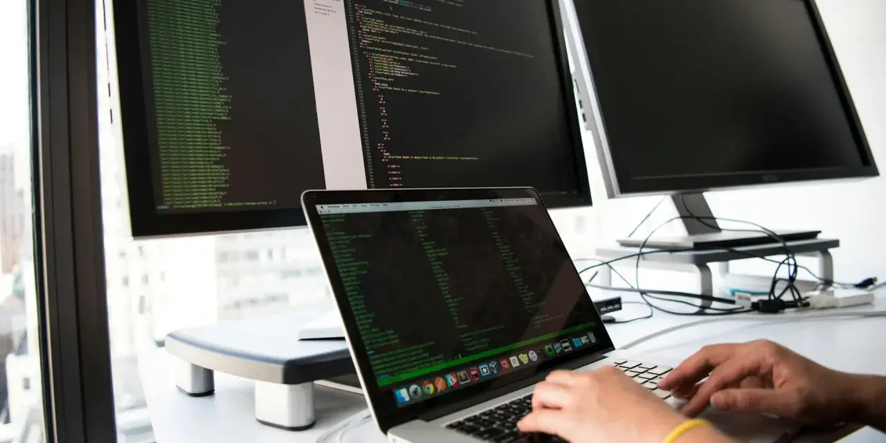 Hands typing on a laptop, surrounded by monitors displaying code. This setup is ideal for developing network infrastructure software.