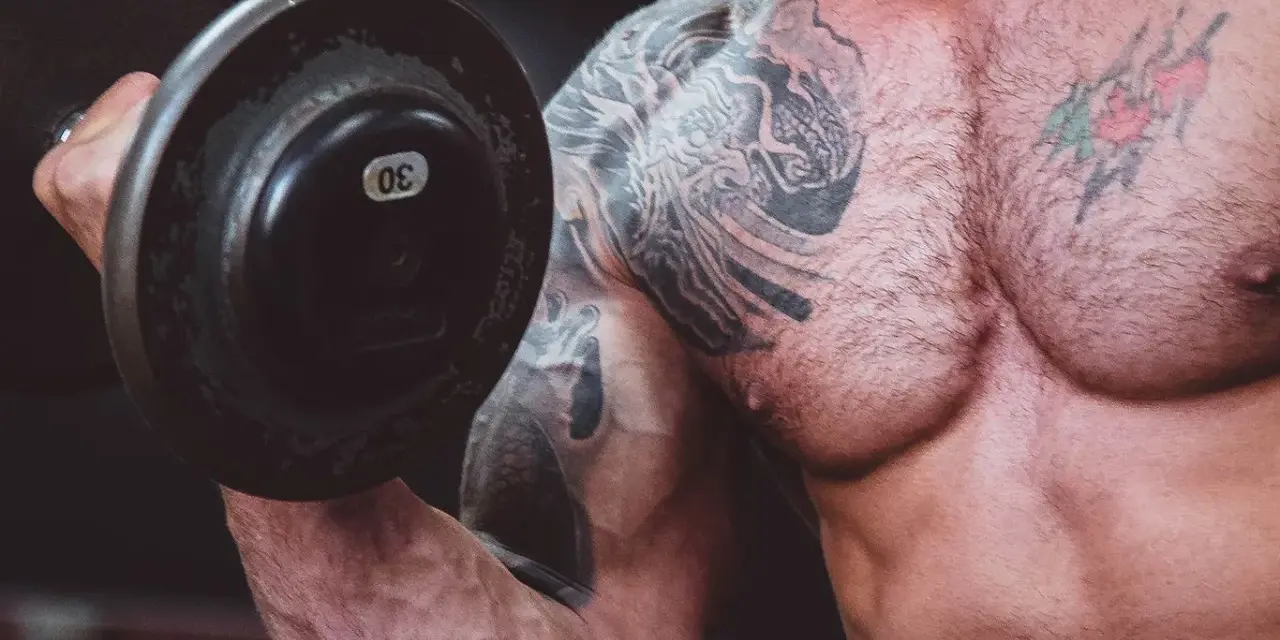 Muskulös man med tatueringar lyfter en 30 kg hantel. Frågan är om man kan bygga muskler på kaloriunderskott.