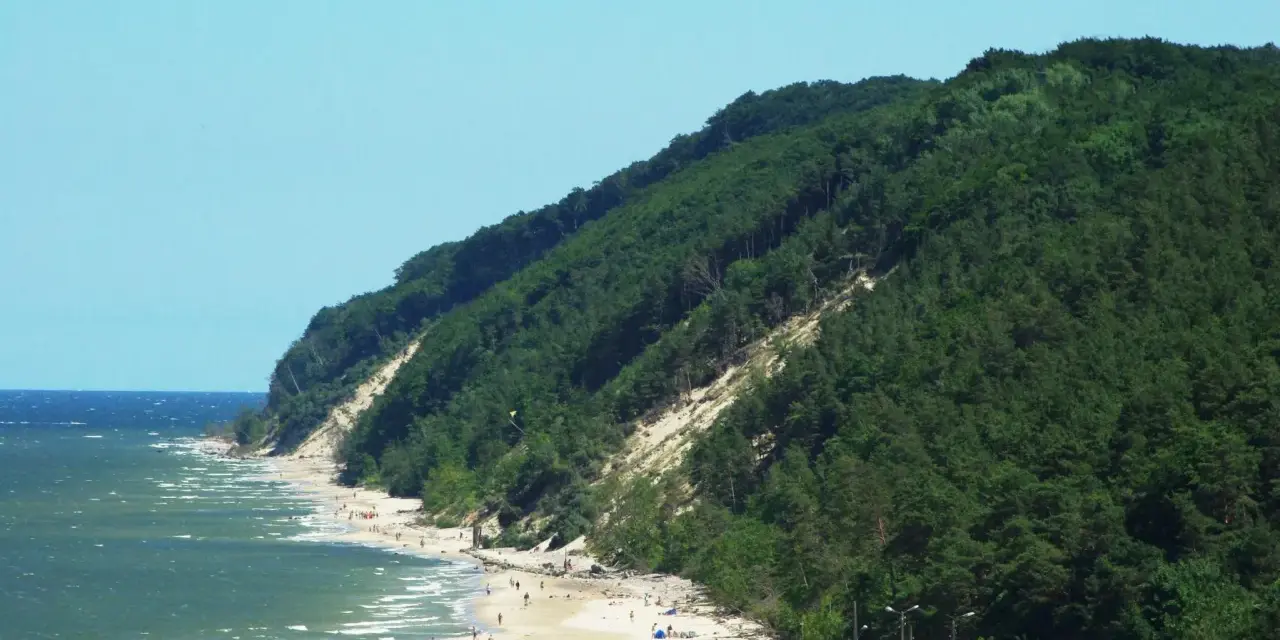 Słoneczna plaża u podnóża zalesionej Kawczej Góry. Woda faluje, a na piasku odpoczywają ludzie.