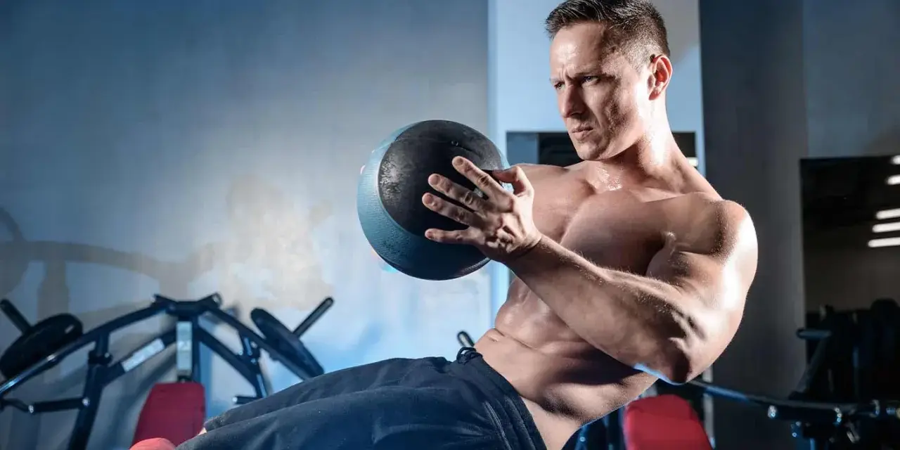 Un homme transpire en faisant un exercice oblique avec un ballon de médecine dans une salle de sport.