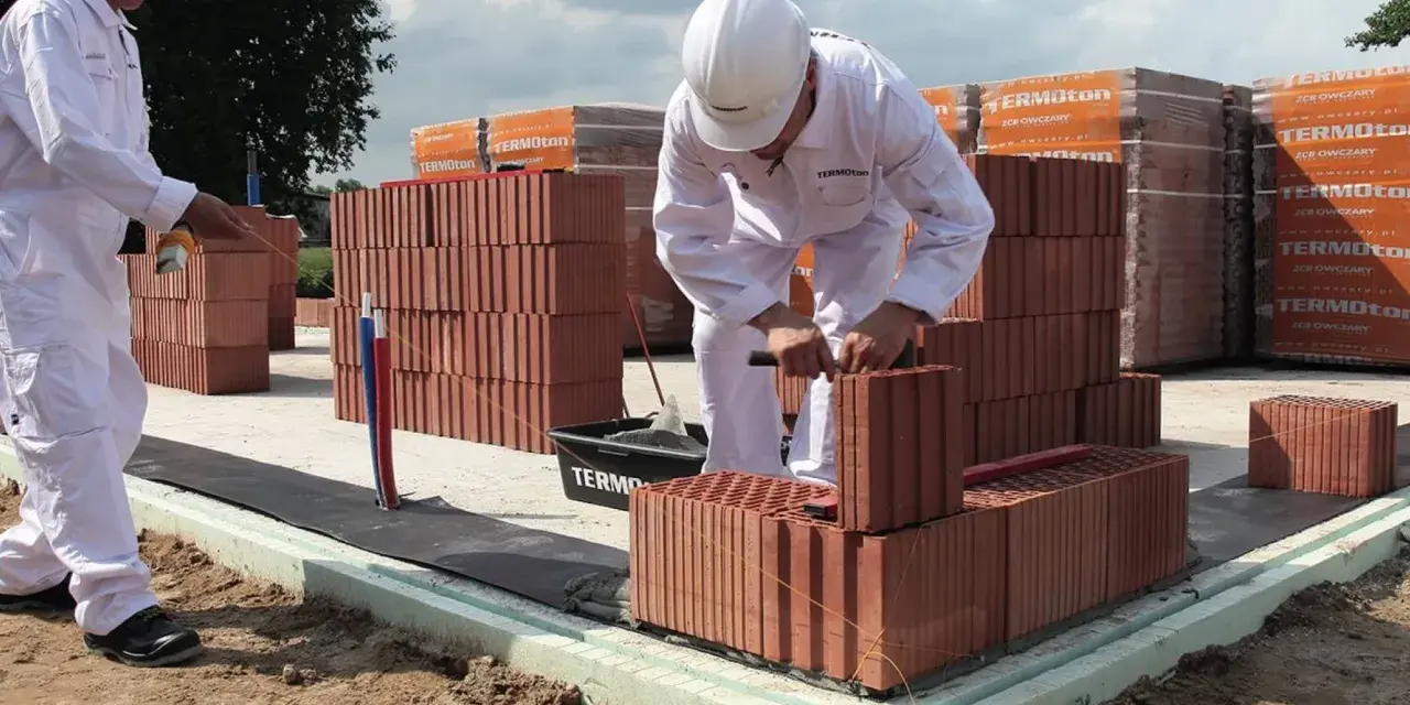 Dwóch budowlańców w białych kombinezonach układa cegły na fundamencie.