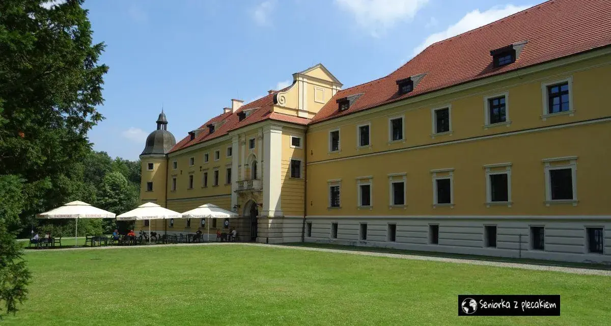 Żółty budynek opactwa cystersów w Rudach z czerwonym dachem i zielonym trawnikiem przed nim.