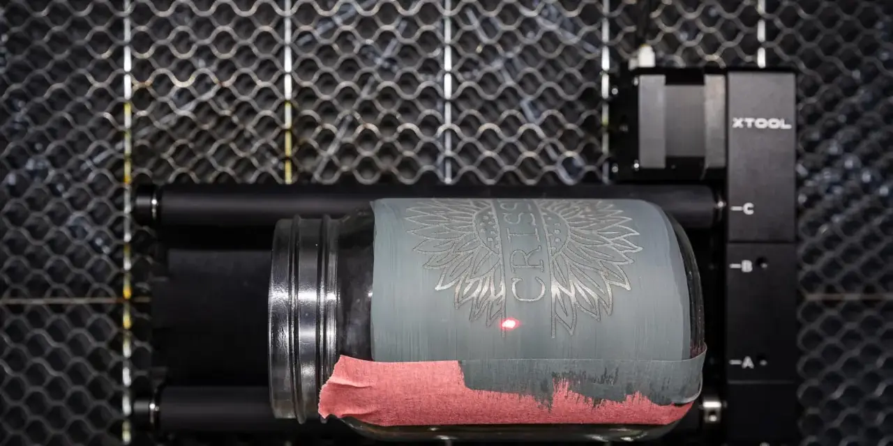 Laser engraving a jar with tempera paint. The laser is creating a sunflower design on the painted glass.