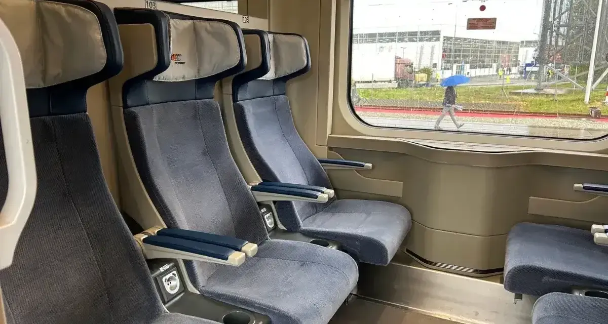 Wygodne siedzenia w wagonie z przedziałami Intercity 2 klasa. Za oknem deszczowa pogoda i przechodzień z niebieskim parasolem.