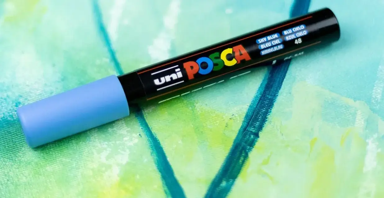 En ljusblå uni POSCA vattenfast tuschpenna ligger på en abstrakt målning i gröna och blå nyanser.