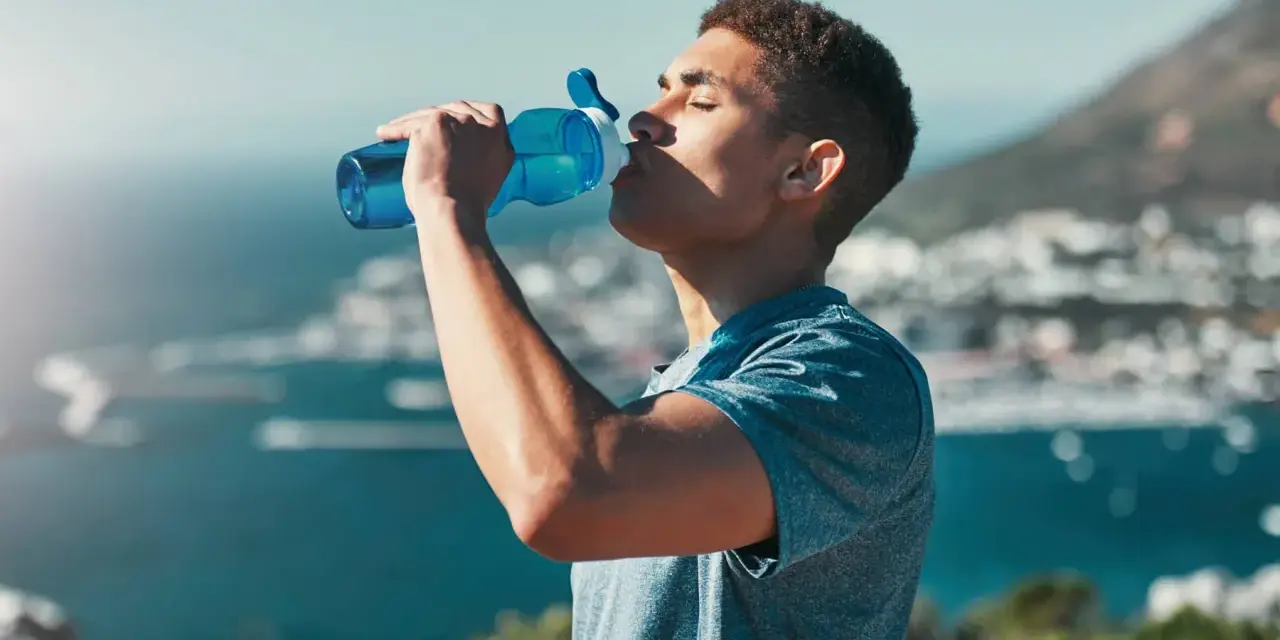 Un jeune homme boit dans une gourde bleue, se réhydratant après l'effort. L'eau, essentielle, contient des électrolytes pour le bon fonctionnement du corps.