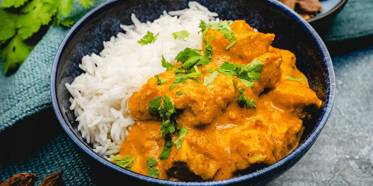Ein cremiges Chicken Korma mit Basmatireis, garniert mit frischem Koriander. Dazu Gewürze und ein kleiner Teller mit Fleischstücken.