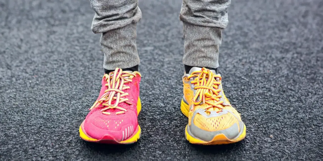 Gambe con pantaloni grigi e scarpe da corsa, una rosa acceso e l'altra arancione, con lacci gialli. La suola scarpa è pronta per correre.