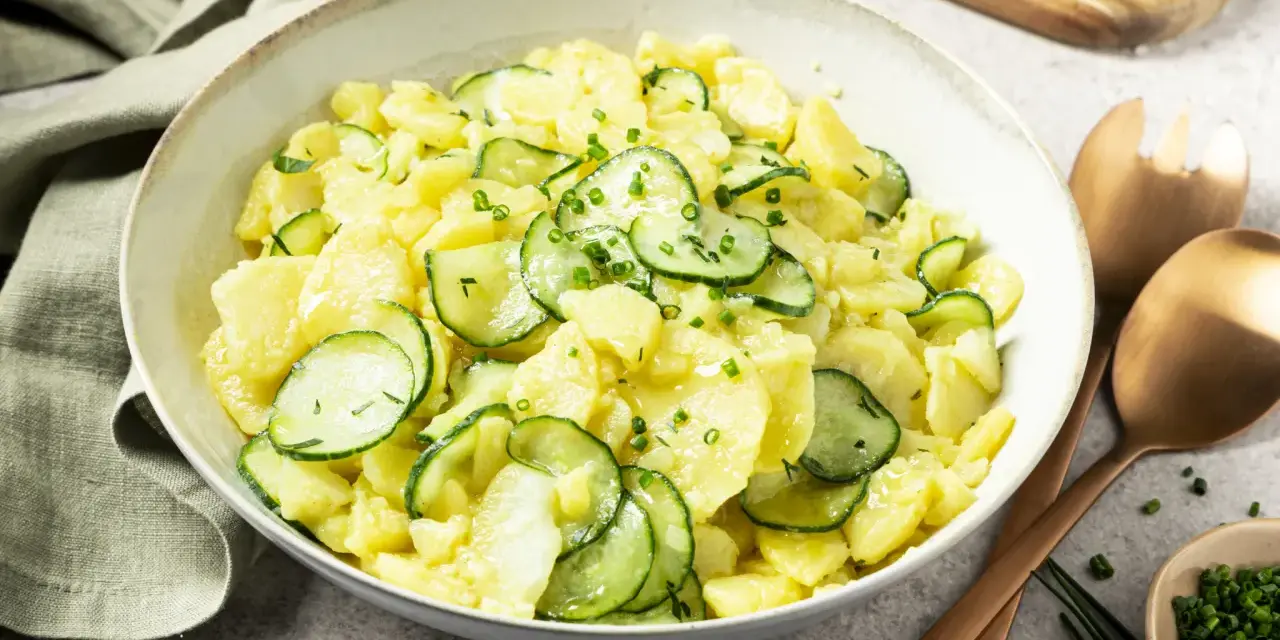 Schüssel mit Kartoffelsalat mit Gurken, garniert mit Schnittlauch. Daneben liegen Servierbesteck und eine kleine Schale mit gehacktem Schnittlauch.
