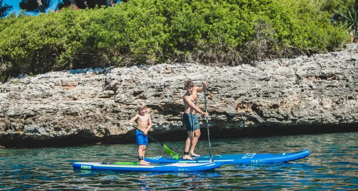 Zwei Jungen auf SUP-Boards, die geradeaus fahren, genießen das klare Wasser vor einer felsigen Küste mit üppiger Vegetation.