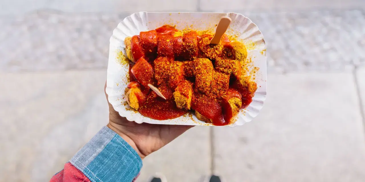 Eine Hand hält eine Portion Currywurst mit original Currywurst Sauce, bestreut mit Currypulver. Ein Holzspieß steckt darin.
