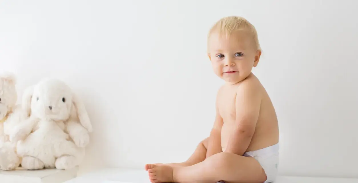 Uśmiechnięty chłopczyk w pieluszce siedzi na wadze. Chyba już czas na większy rozmiar, kiedy pampers jest za mały.
