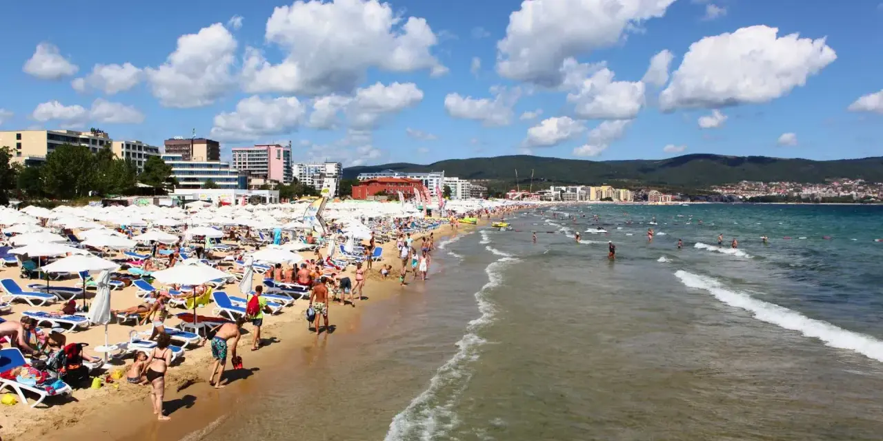 Tłum ludzi wypoczywa na piaszczystej plaży Słoneczny Brzeg, opalając się na leżakach pod białymi parasolami. W tle widać morze i zabudowania kurortu.