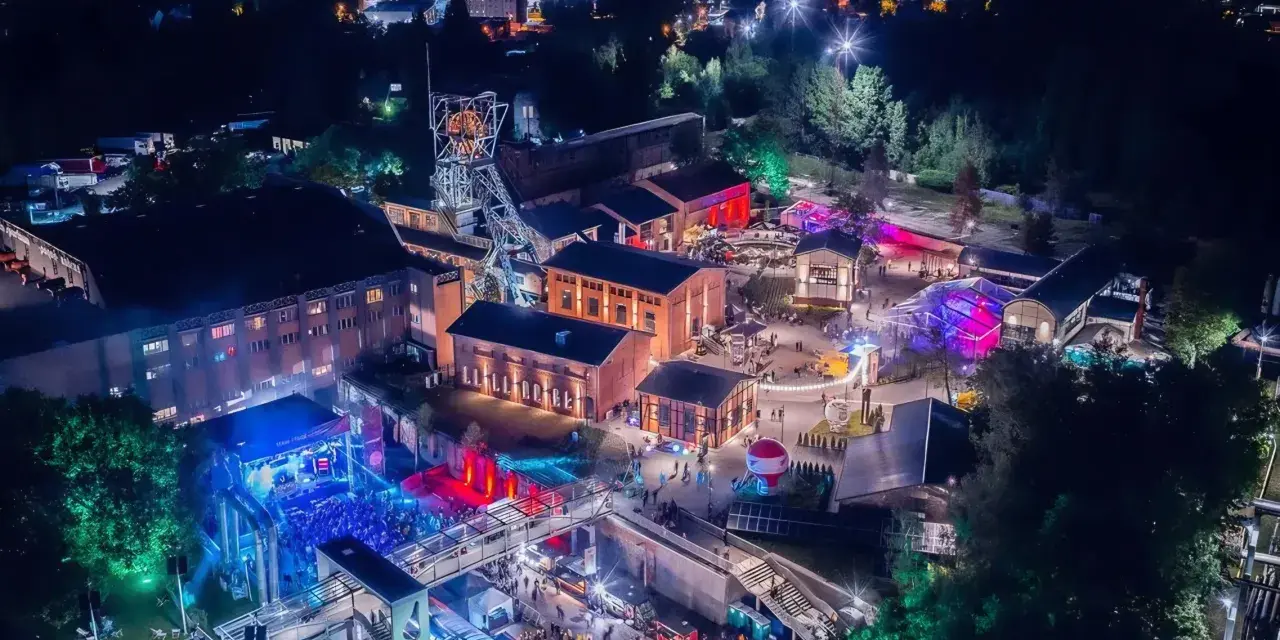 Nocny festiwal Carbon Silesia rozświetla postindustrialny krajobraz. Tłumy bawią się przy scenie, a kolorowe światła odbijają się od budynków dawnej kopalni.