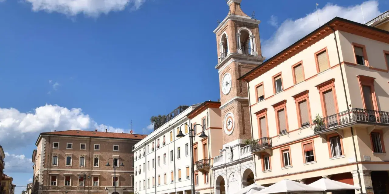 Piazza Tre Martiri a Rimini: un campanile con orologio domina la scena, tra palazzi storici e tavolini all'aperto. Ecco cosa vedere!