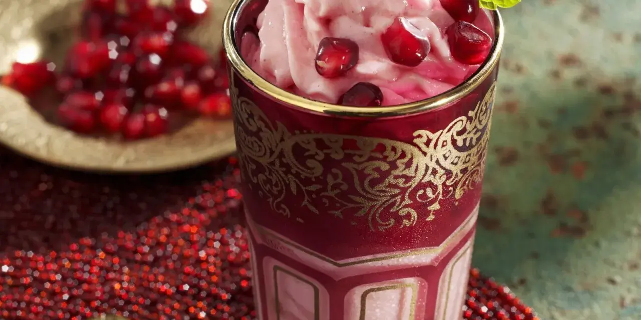 Ein Glas mit cremigem granatapfel eis, garniert mit frischen Granatapfelkernen und Minze. Daneben eine Schale mit Granatapfelkernen.