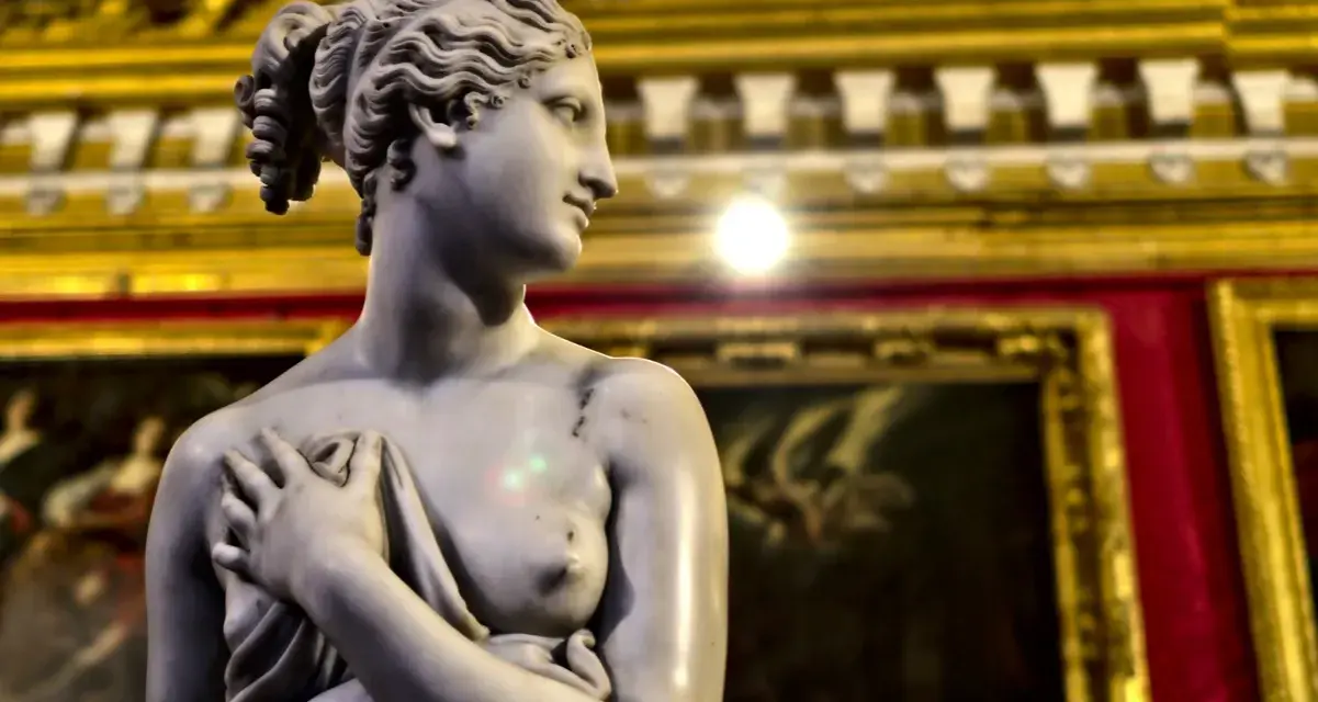 Rzeźba Wenus, dzieło Antonio Corradini, z dłonią na piersi, na tle złoconych ram obrazów.