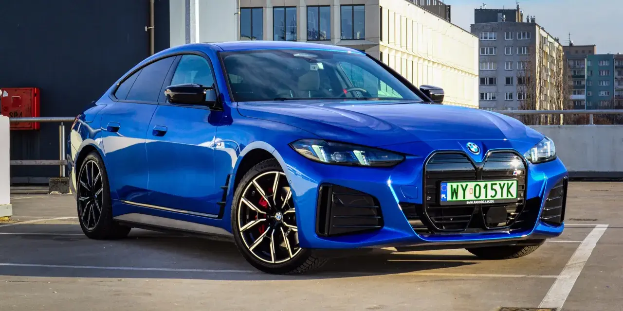 Niebieskie BMW i4 M50 zaparkowane na parkingu, z widokiem na budynki w tle.