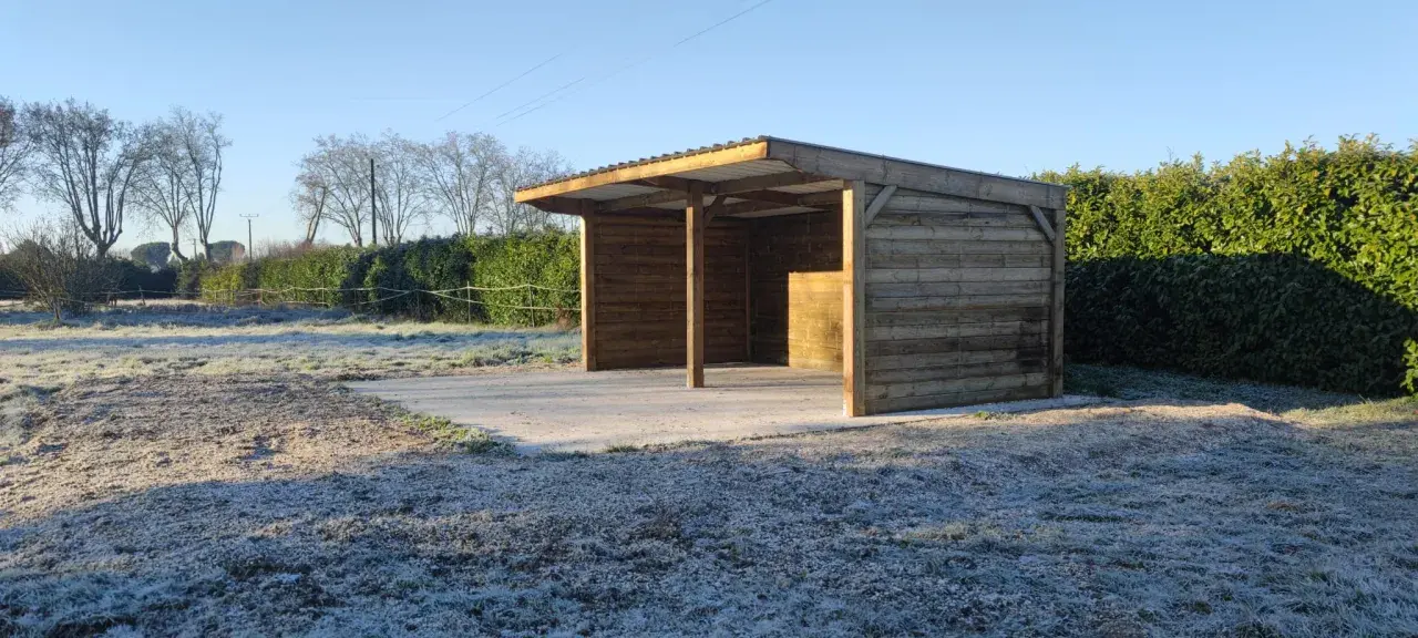 Abri pour chevaux d'occasion en bois, avec toit métallique, sur un sol gelé. Parfait pour protéger vos équidés du froid.